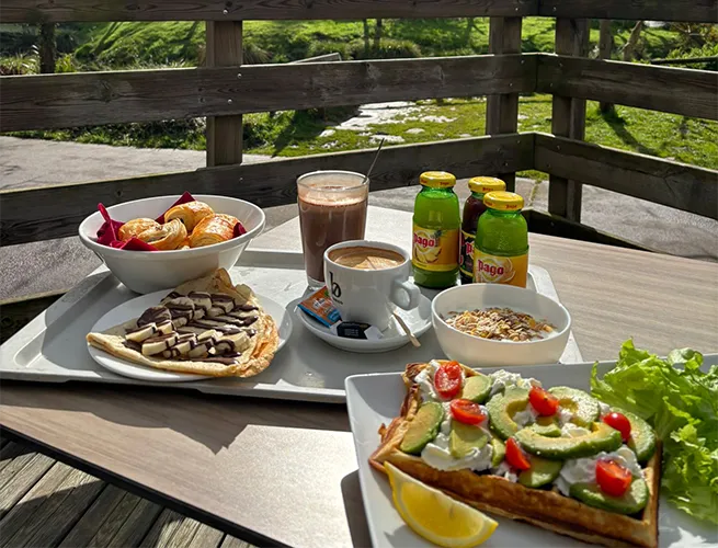 Plateau de brunch sur une terrasse en bois : tartine à l'avocat, viennoiseries, chocolat chaud et jus