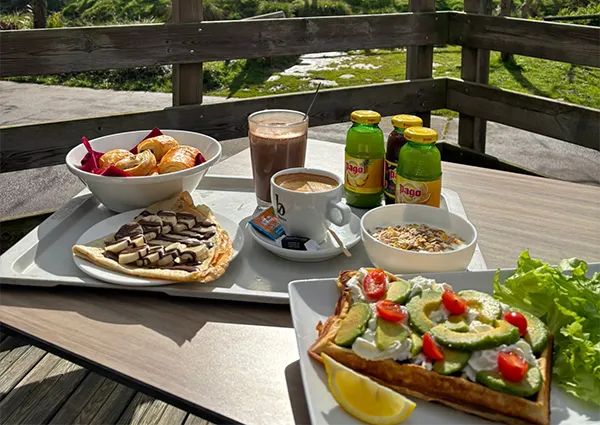 Plateau de brunch sur une terrasse en bois avec tartine à l'avocat, crêpe, chocolat chaud et jus